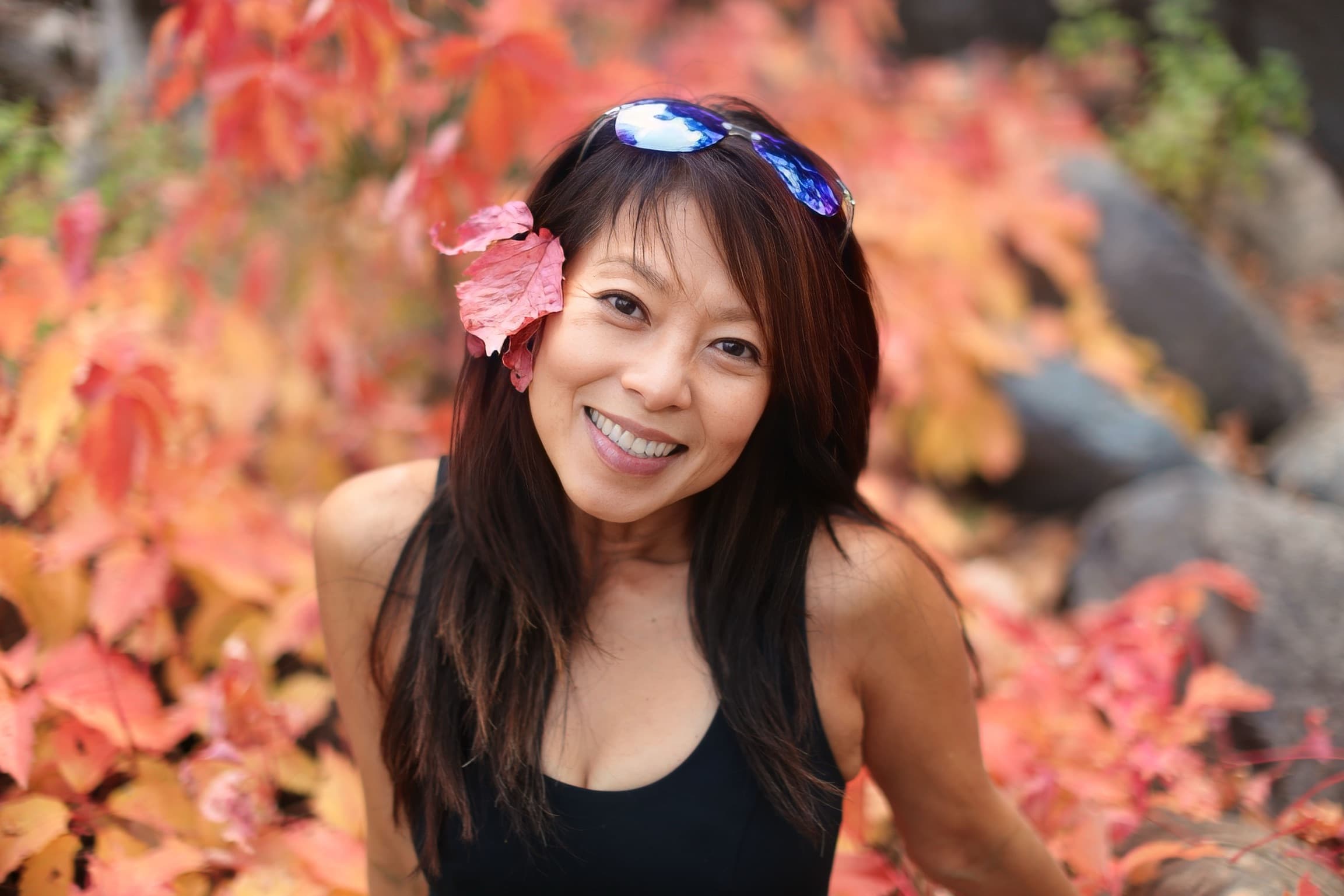 Nicole Pham smiling among bright autumn leaves, sunglasses on her head and a red leaf tucked behind her ear